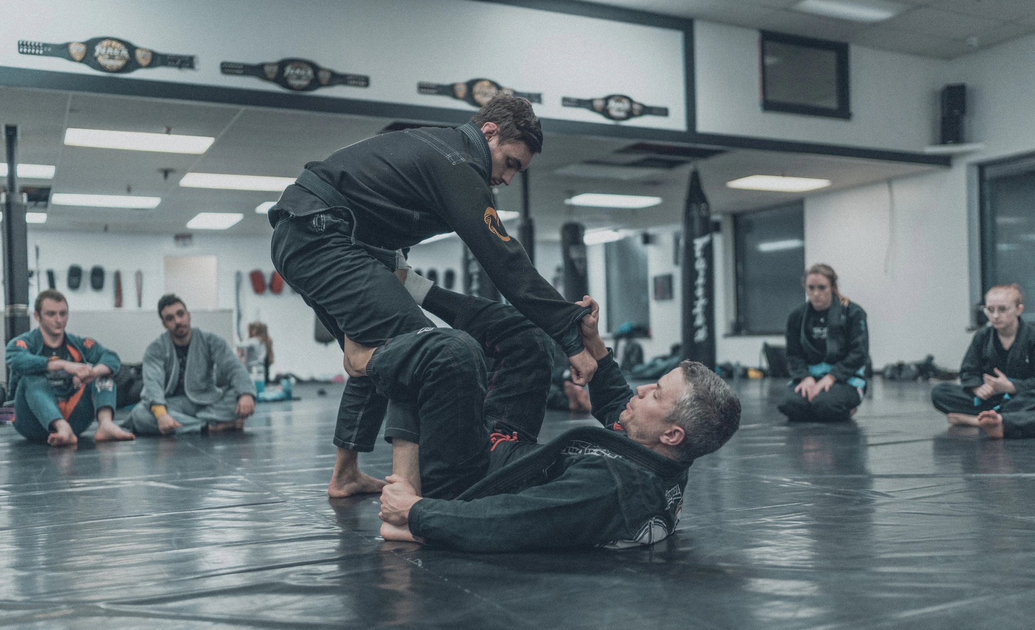 Adult Brazilian Jiu-Jitsu rolling on the mat at Precision — The Barracks, Collegeville PA — championship belts visible on the wall